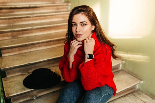 Chica de pelo oscuro en suéter rojo se sienta en las escaleras. Foto de mujer con ojos marrones