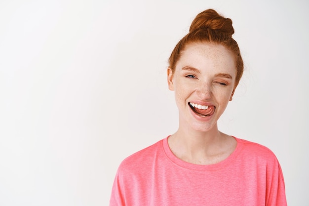 Chica con el pelo pelirrojo peinado en un moño desordenado, guiñando un ojo con alegría y mostrando una sonrisa perfectamente blanca, sacando la lengua después de cepillarse los dientes, de pie contra la pared blanca