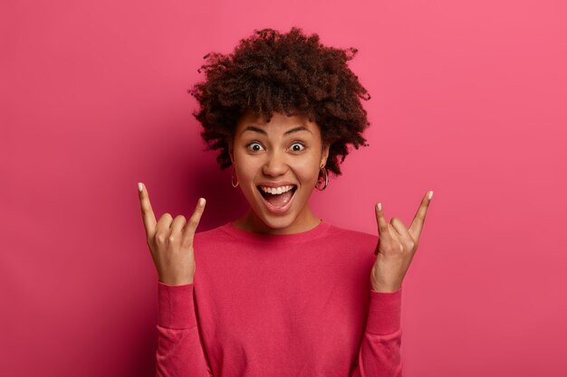 Chica de pelo rizado positiva hace gesto de cuerno, disfruta de la música rock n roll, se ríe de alegría, usa un jersey informal