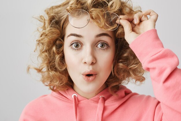 Chica de pelo rizado sorprendida se quita las gafas y dice wow asombrada