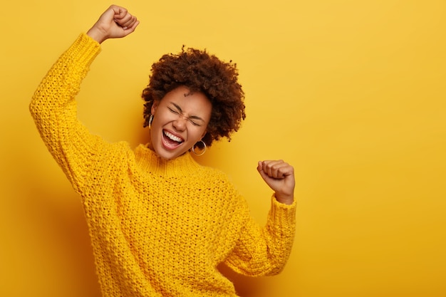 Chica de pelo rizado en suéter amarillo de invierno baila con los brazos extendidos en el aire, disfruta de la música, tiene expresión de la cara llena de alegría, posa en interiores.