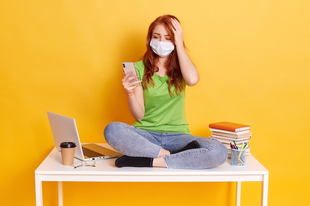 Chica de pelo rojo desconcertado sentado en la mesa con el teléfono en las manos leyendo noticias impactantes en el trabajo de la red social, manteniendo la mano en la cabeza