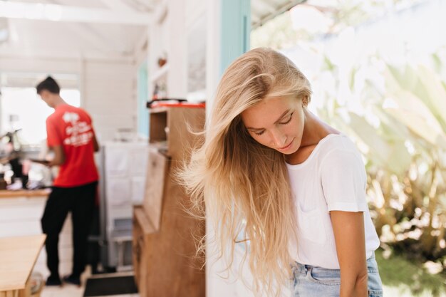 Chica pensativa con cabello largo claro posando en casa. Filmación en interiores de modelo femenino blanco de ensueño en camiseta de moda mirando hacia abajo mientras está de pie en la cafetería.