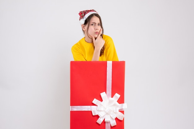 Chica pensativa con gorro de Papá Noel de pie detrás de un gran regalo de Navidad en blanco