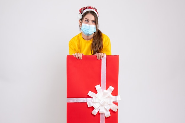 Chica pensativa con gorro de Papá Noel de pie detrás de un gran regalo de Navidad en blanco