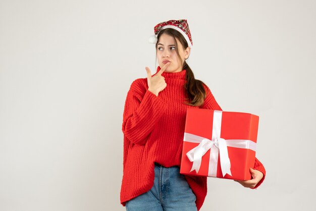 Chica pensativa con gorro de Papá Noel sosteniendo presente poniendo el dedo en los labios de pie sobre blanco