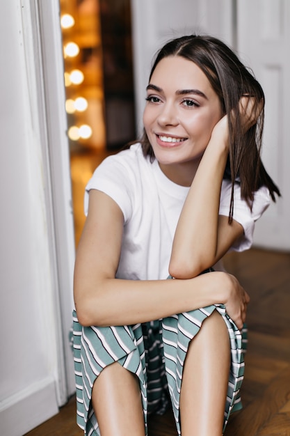 Chica pensativa con grandes ojos oscuros sentada en el suelo. Bella dama sonriente en pijama pasando el fin de semana en su apartamento.