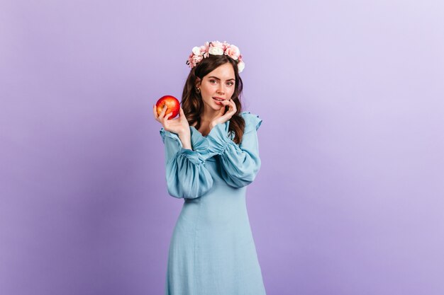 Chica pensativa mirando con picardía, sosteniendo apetitosa manzana. Retrato de modelo con corona de flores en pared lila.