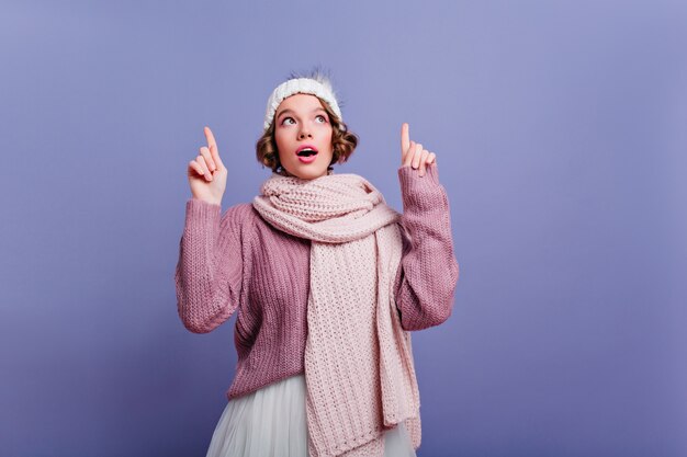 Chica pensativa de pelo corto con sombrero lindo mirando hacia arriba con la boca abierta. Modelo femenino despreocupado posando en accesorios de invierno en la pared púrpura.