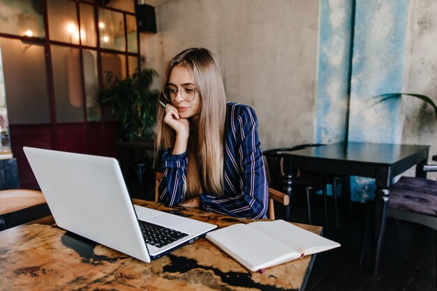 Chica pensativa de pelo largo con gafas mirando la pantalla del portátil. Encantadora mujer morena sentada en la cafetería con la computadora.
