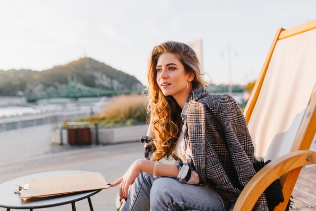 Chica pensativa de pelo largo en jeans esperando su pedido en la cafetería al aire libre