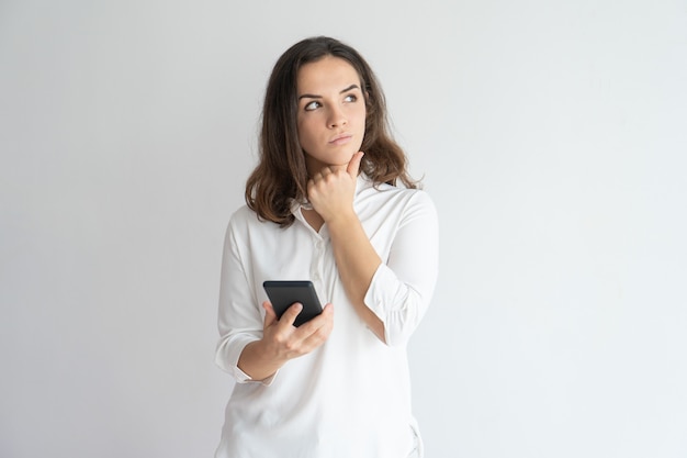 Chica pensativa pensando en mensaje de texto. Joven mujer caucásica sosteniendo teléfono inteligente