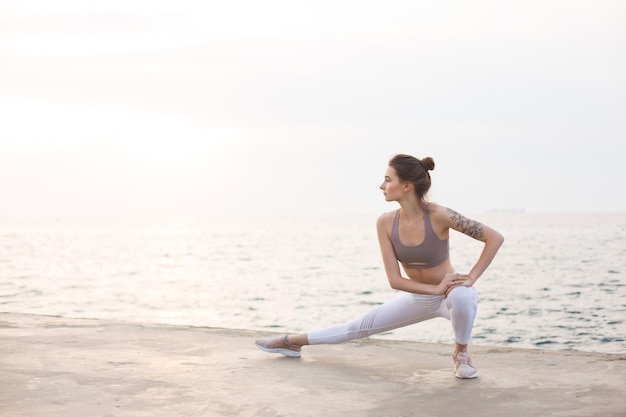 Chica pensativa en top deportivo y polainas blancas practicando yoga con increíbles vistas al mar en el fondo. mujer joven, extensión, por, el, mar