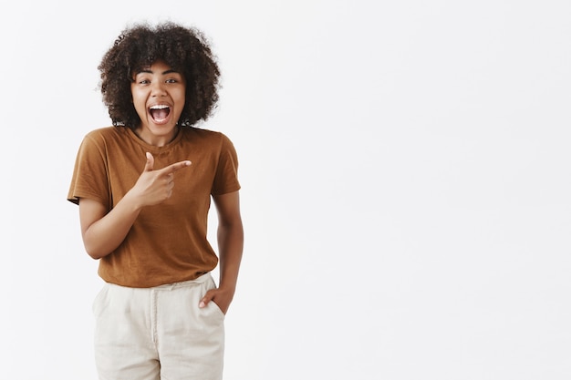 Chica de piel oscura con peinado afro en elegante camiseta marrón divirtiéndose pasando tiempo riendo alegremente y señalando algo divertido a la derecha tomados de la mano en el bolsillo