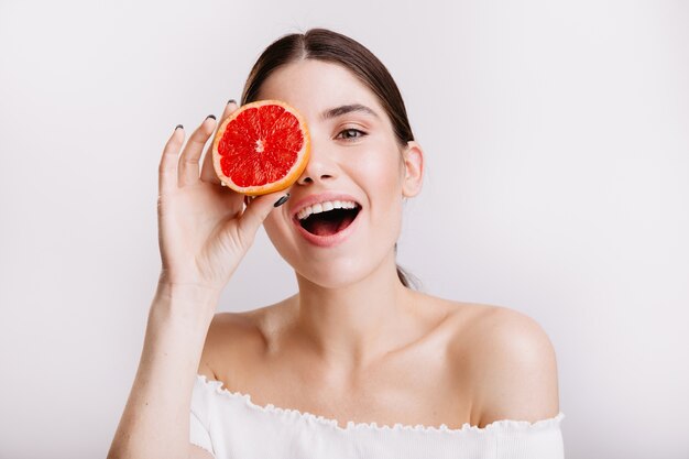 Chica con piel perfecta posa felizmente en pared aislada. Retrato de modelo femenino apoyando una dieta saludable.