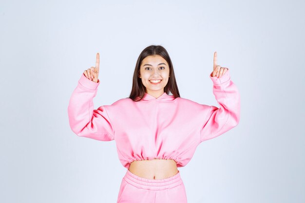 Chica en pijama rosa apuntando al piso de arriba con cara emocional