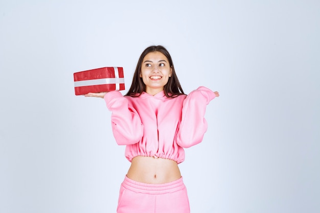 Foto gratuita chica en pijama rosa sosteniendo una caja de regalo rectangular roja y parece satisfecha.