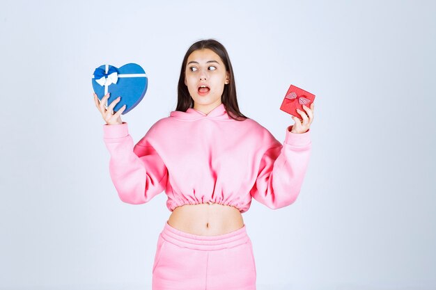Chica en pijama rosa sosteniendo cajas de regalo con forma de corazón rojo y azul y haciendo una elección entre ellos.