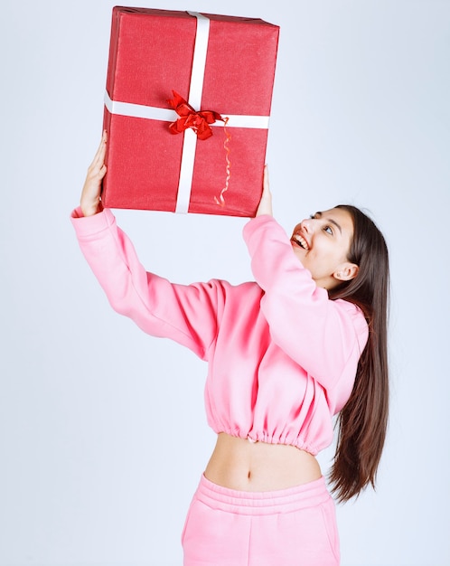 Chica en pijama rosa sosteniendo una gran caja de regalo roja sobre su cabeza.