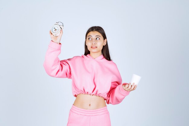 Chica en pijama rosa sosteniendo un reloj despertador y una taza de café con cara cansada.