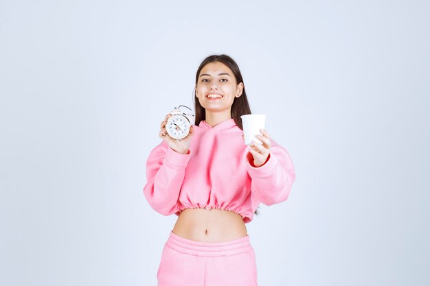 Chica en pijama rosa sosteniendo un reloj despertador y una taza de café.