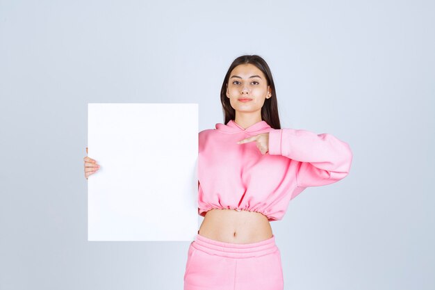 Chica en pijama rosa sosteniendo un tablero de presentación cuadrado en blanco y apuntando a él.