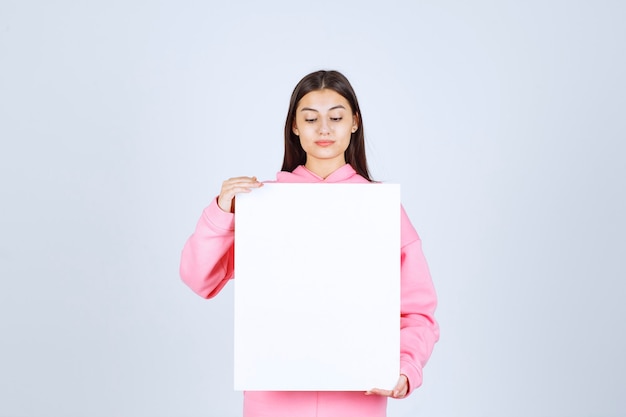 Chica en pijama rosa sosteniendo un tablero de presentación cuadrado en blanco frente a ella.