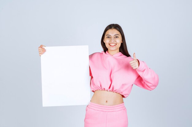 Chica en pijama rosa sosteniendo un tablero de presentación cuadrado en blanco y mostrando el pulgar hacia arriba.