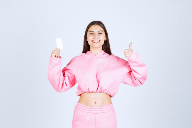 Chica en pijama rosa sosteniendo una tarjeta de visita y mostrando signo de mano de disfrute.