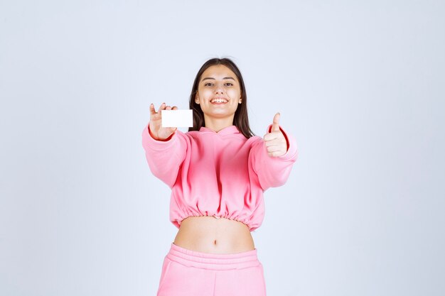 Chica en pijama rosa sosteniendo una tarjeta de visita y mostrando signo de mano de disfrute.