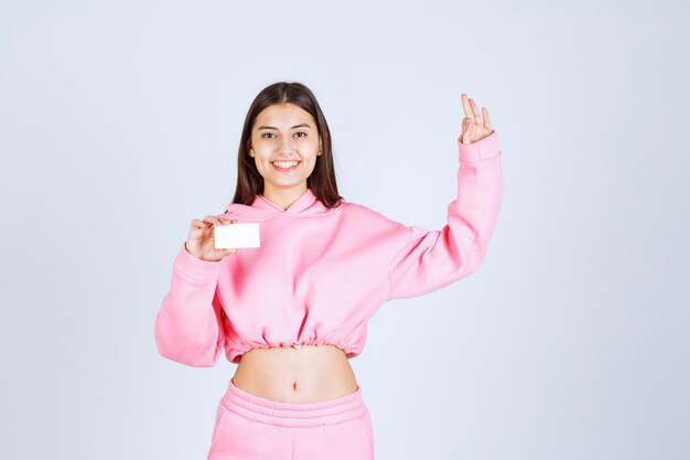 Chica en pijama rosa sosteniendo una tarjeta de visita y mostrando signo de mano de disfrute.
