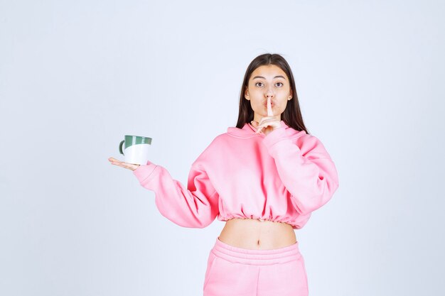 Chica en pijama rosa sosteniendo una taza de café y pidiendo silencio.