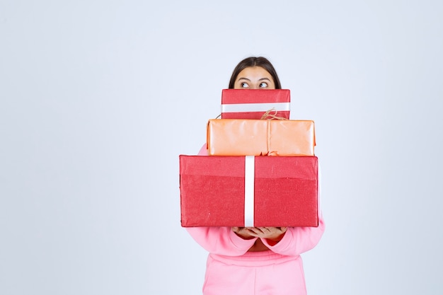 Chica en pijama rosa sosteniendo varias cajas de regalo rojas y escondiendo su rostro detrás de ellas.