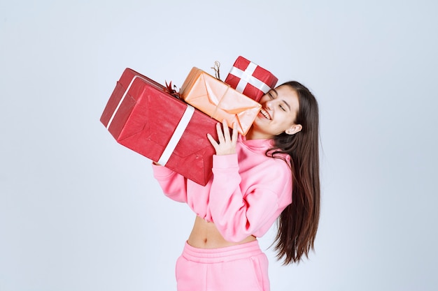 Chica en pijama rosa sosteniendo varias cajas de regalo rojas y sintiéndose feliz.