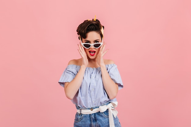 Chica pinup alegre posando con gafas de sol. Disparo de estudio de mujer joven en blusa de rayas azules.