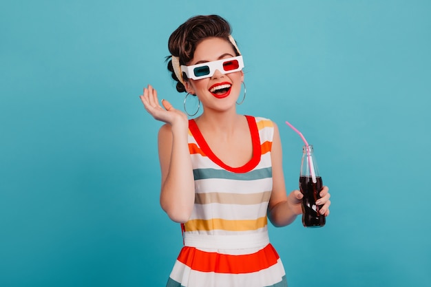 Chica pinup de buen humor posando en gafas 3d. Disparo de estudio de mujer morena dichosa con botella de refresco aislado sobre fondo azul.