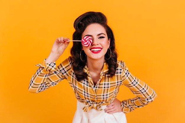 Chica pinup despreocupada con piruleta. Foto de estudio de risa elegante mujer en camisa a cuadros divirtiéndose sobre fondo amarillo.