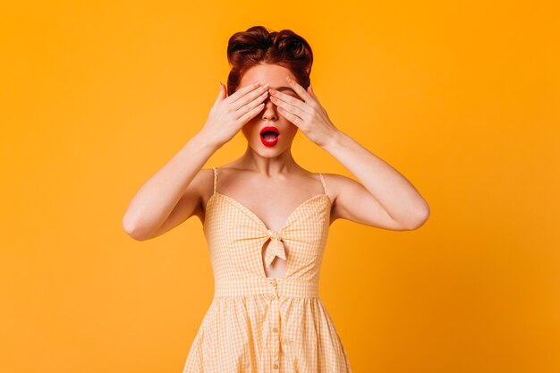 Chica pinup divertida que cubre los ojos con las manos. Disparo de estudio de increíble mujer de jengibre en vestido aislado en espacio amarillo.