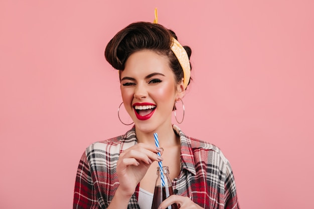 Chica pinup emocional bebiendo bebidas. Disparo de estudio de mujer joven emocionada en camisa a cuadros aislada sobre fondo rosa.