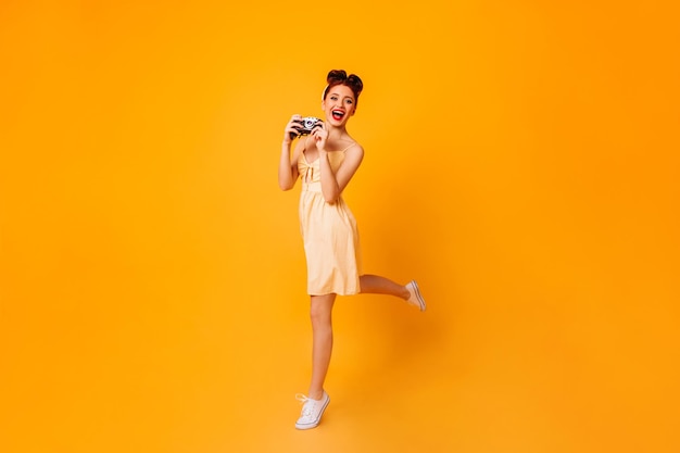 Chica pinup emocional con cámara bailando sobre fondo amarillo Foto de estudio de fotógrafa vestida