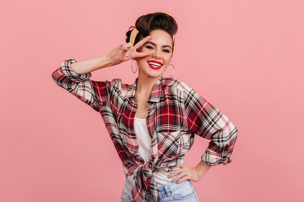 Chica pinup feliz posando con el signo de la paz. Foto de estudio de risa a mujer hermosa en camisa a cuadros roja.