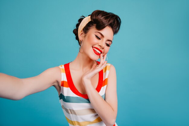 Chica pinup refinada sonriendo con los ojos cerrados. Jovencita morena de moda tomando selfie sobre fondo azul.