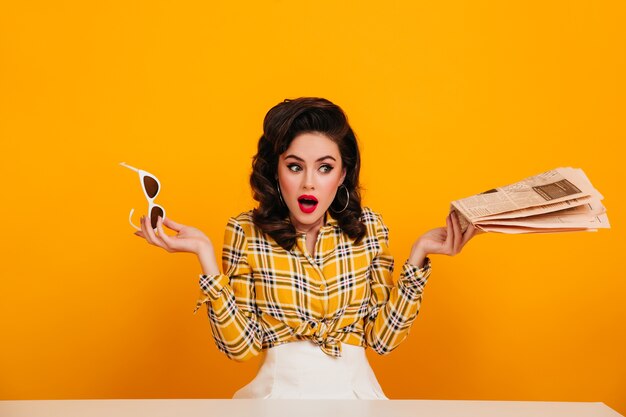 Chica pinup sorprendida con gafas de sol sobre fondo amarillo. Mujer joven elegante posando con periódico.