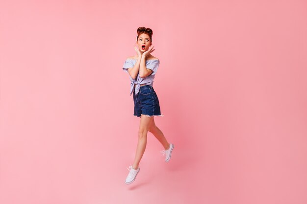 Chica pinup sorprendida en zapatos blancos posando con la boca abierta. Vista de longitud completa de la mujer de jengibre emocional saltando en el espacio rosa.