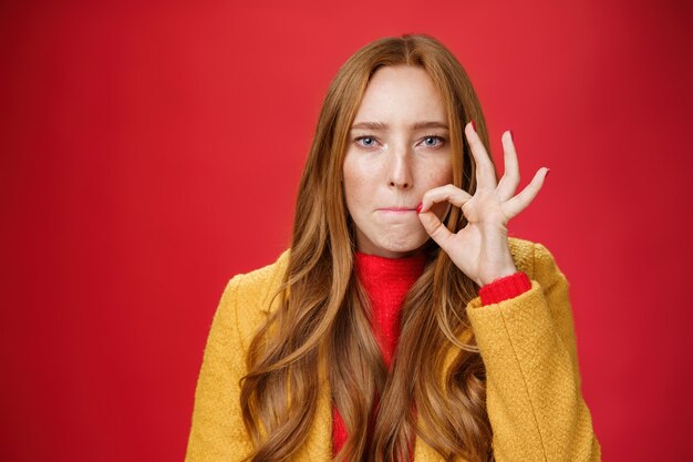 Chica poniendo sello en la boca haciendo la promesa de no contarle a nadie secretos, chupándose los labios y sosteniendo el dedo cerca para no derramar palabras, luciendo seria y decidida a mantener la sorpresa a salvo sobre la pared roja.