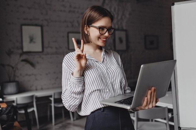 Chica positiva en auriculares y gafas muestra el signo de la paz, hablando por video en la computadora portátil en el lugar de trabajo.