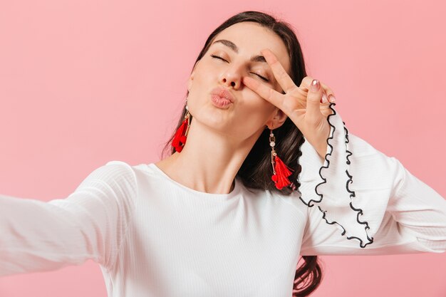 Chica positiva en blusa blanca toma selfie y muestra el signo de la paz sobre fondo rosa.