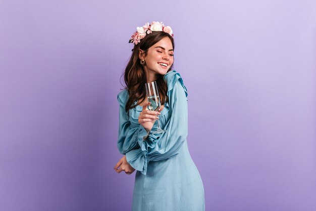 Chica positiva de buen humor se ríe mientras disfruta de la fiesta en la pared púrpura. Modelo en traje azul con copa de champán.
