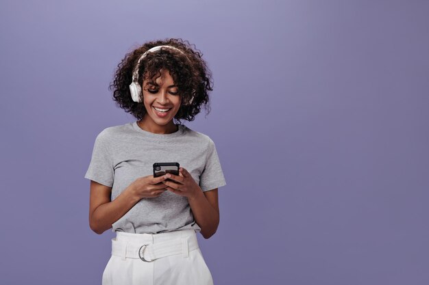 Chica positiva en camiseta brillante con una sonrisa hablando por teléfono y escuchando música Mujer elegante en camiseta gris y pantalones cortos blancos disfrutando de canciones en auriculares