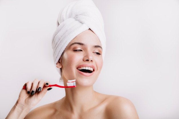 Chica positiva sin maquillaje lindo sonríe en la pared blanca. Mujer después de la ducha cepillándose los dientes.